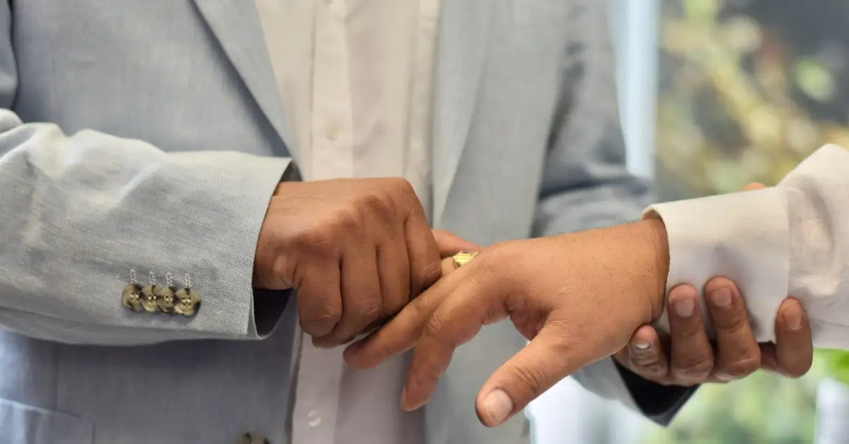 Una mano de hombre sostiene otra y le coloca un anillo en un dedo. Uno viste traje azul y el otro, blanco.jpg