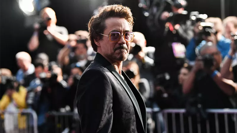 Foto de Robert Downey Jr. con saco negro, lentes cuadrados y barba, con fotógrafos detrás de él.