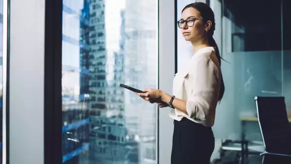 Mujer dentro de su oficina sosteniendo una tablet y viendo hacia el horizonte.