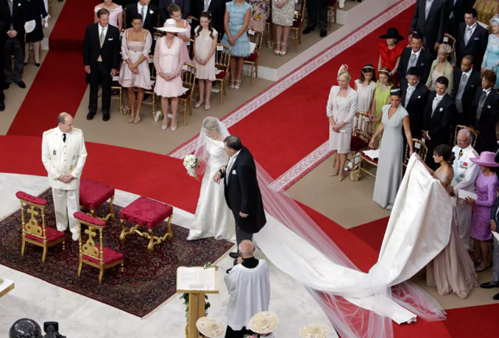 Charlene optó por la sencillez. No porta joyas y tampoco tiara. La cola del vestido es muy larga y el velo también es sencillo. Podemos decir que Charlene es una novia clásica. Camina de la mano de Michael, su padre.