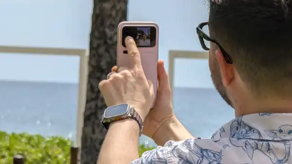 Una persona vista de espaldas sostiene un teléfono rosa frente al mar y toca la pequeña pantalla situada junto a las cámaras traseras. En la pantalla aparece la vista previa de una fotografía.