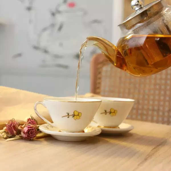 Foto de una tetera de vidrio transparente sirviendo té en dos tazas de cerámica con orillas doradas y dibujos flores amarillas.