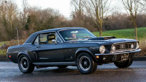 Un Ford Mustang clásico negro de 1970 —un automóvil estadounidense— circulando por una carretera rural inglesa.