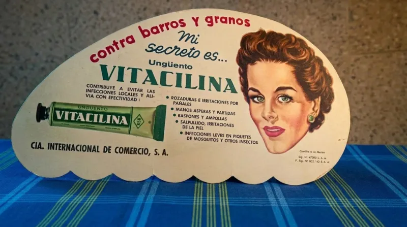 Anuncio publicitario en forma de abanico de Vitacilina. Es de aspecto medio ovalado y en él se ve el rostro de una mujer en los 60, un tubo de Vitacilina y frases como "Mi secreto es..." y el nombre de la marca. 