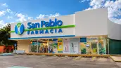 Una sucursal de San Pablo  Farmacia  vista desde el estacionamiento, con una fachada blanca y moderna que muestra el logotipo azul y verde de San Pablo y el nombre de farmacia en letras grandes. Al frente se observan los accesos de la tienda y los ventanales con anuncios de productos y servicios, además de un área identificada como San Pablo Clínica. El establecimiento cuenta con espacios de estacionamiento y señalización para personas con discapacidad. La escena está iluminada por luz natural, con árboles alrededor y un cielo azul con nubes blancas al fondo.