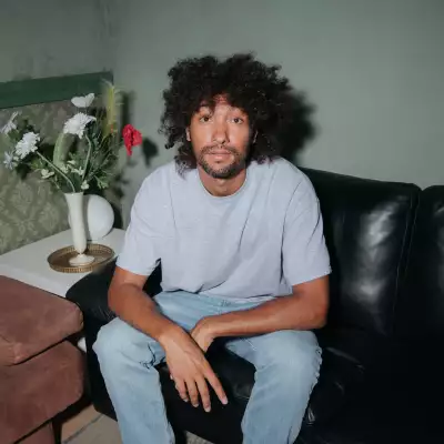 Foto del DJ &Me, sentado en una sala de piel negra, en una habitación de paredes verdes, él viste playera gris y jeans.