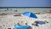 Playa de Cancún con arena blanca, mar turquesa y turistas disfrutando del agua; en primer plano hay una sombrilla azul, una colchoneta inflable y toallas sobre la arena.