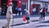 Dos personas de la tercera edad caminan apoyados en un bastón y una andadera en una calle del centro de Cuernavaca.