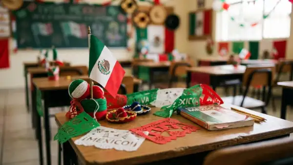 Un pupitre con decoraciones de México por la independencia del país, sobre él hay una bandera miniatura de México, papel picado color verde, blanco y rojo, y dos pequeños sombreros de mariachi, en un salón de clases también decorado. 