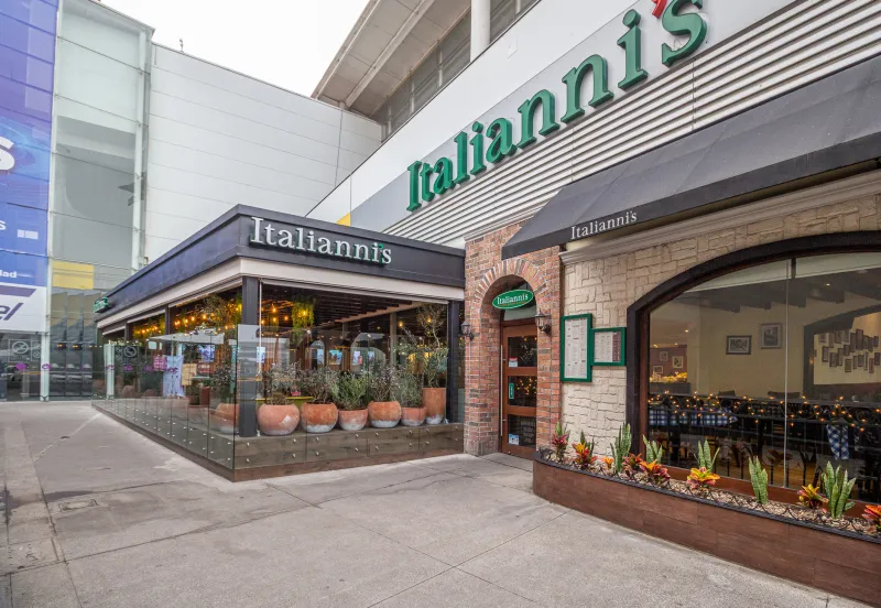 :Fachada de un restaurante Italianni’s en un centro comercial, con ventanales amplios, jardineras, detalles de ladrillo y piedra, y letreros con el nombre de la marca.