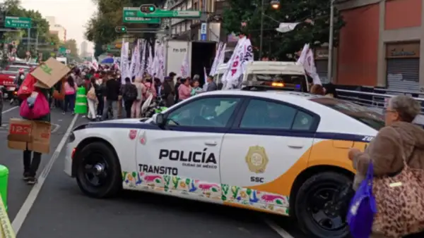 Una patrulla color blanco con vivos amarillos con letras que dice policía y tránsito en una calle con personas y pancartas.jpg