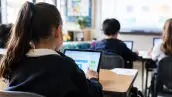 Niños de espaldas utilizan tabletas electrónicas en un salón de clases mientras realizan actividades escolares.