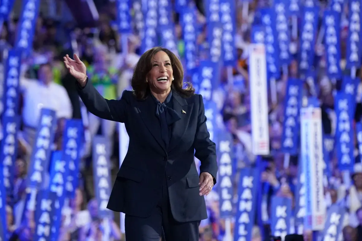 Kamala Harris, una mujer negra de 59 años sonriendo. Usa un traje azul marino saludando al público.