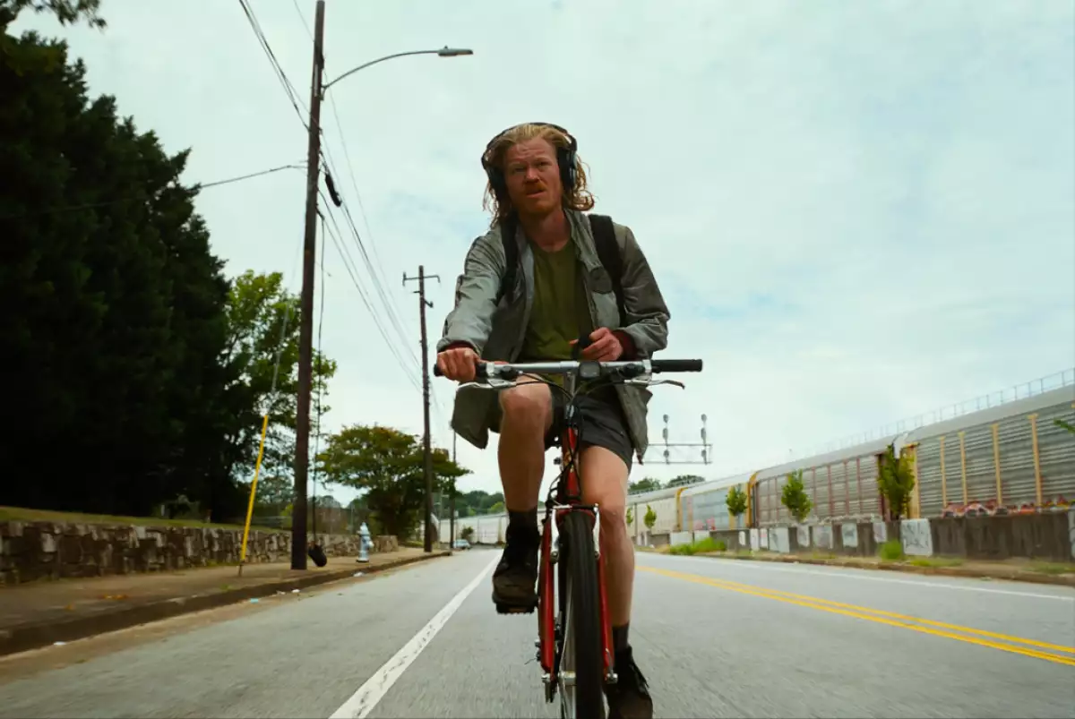 Foto de Jesse Plemons en bicicleta vestido de shorts, cabello largo y con audífonos de diadema en la película 'Bugonia'.
