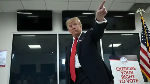 Donald Trump, con traje oscuro y corbata roja, levanta el brazo y señala hacia arriba dentro de un centro de votación en Florida. A su lado hay un cartel que invita a ejercer el derecho al voto y una bandera de Estados Unidos.