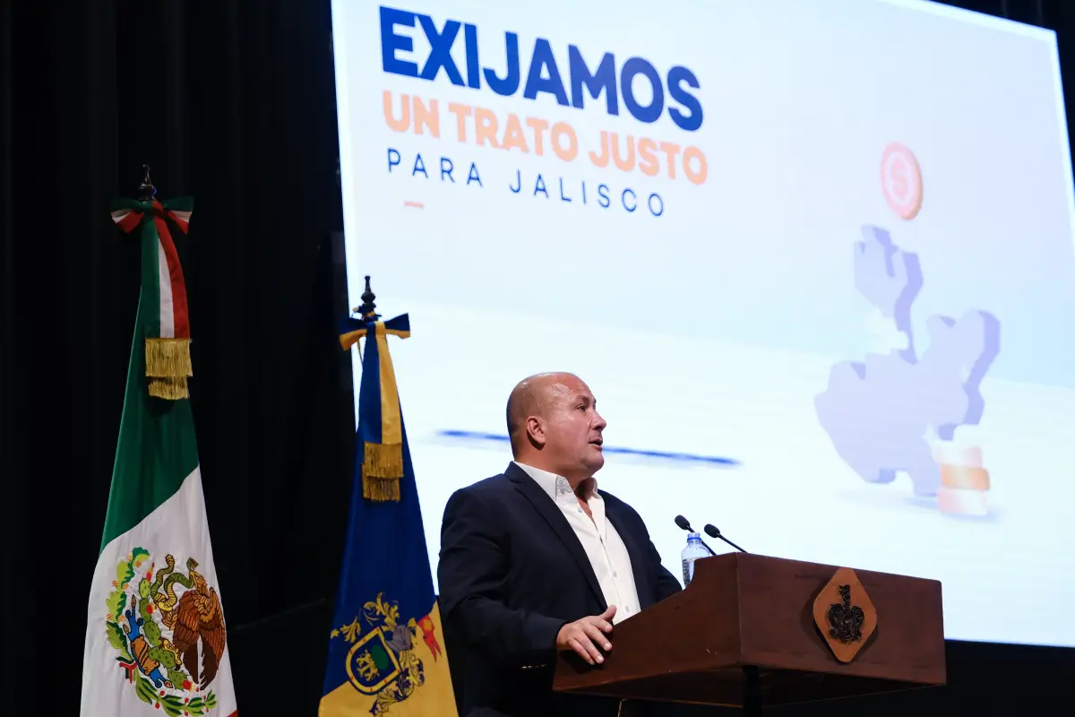 El gobernador, Enrique Alfaro, habla parado en un podio donde a su lado derecho está la bandera de México y la Jalisco, a su espalda hay una pantalla  grande donde de lee: "Exijamos trato justo para Jalisco".