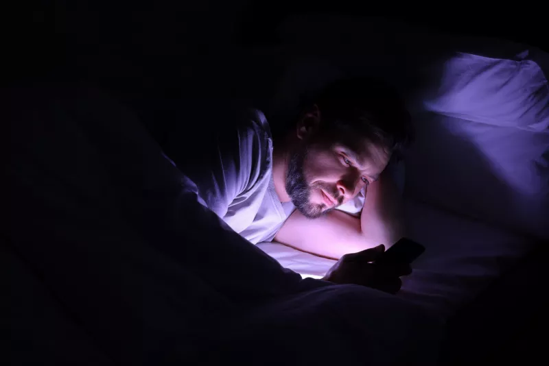 Foto de stock de un hombre acostado en su cama. en un cuarto completamente oscuro y únicamente ilumina su cara el celular