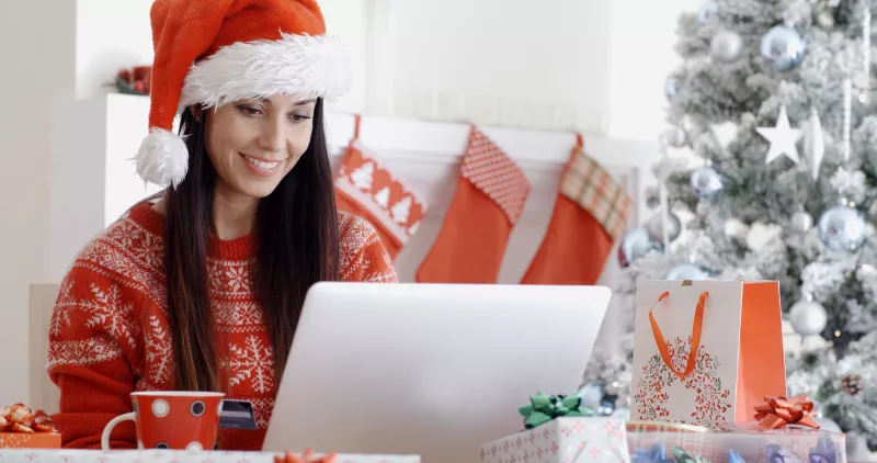 Mujer haciendo compras en una laptop. Trae un gorro de navidad y un suéter navideño. Alrededor de ella hay cajas de regalos, una taza de café y al fondo, un árbol de Navidad.