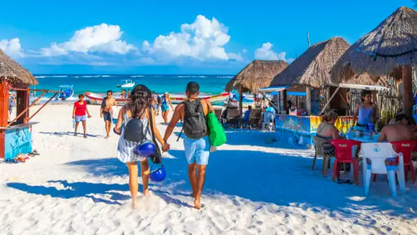 Una pareja camina de mano en una playa con arena blanca y mar y cielo azul. Palapas y sillas donde hay personas sentadas en sillas.jpg