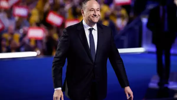 Doug Emhoff, un hombre de 59 años vistiendo un traje negro, camisa blanca y corbata azul. Va caminando sobre un escenario con el fondo de cientas de personas y carteles rojos con su nombre.