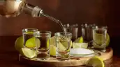 Seis caballitos de tequila sobre una plataforma circular de madera. Una mano sirve tequila blanco. En la tabla redonda también hay limones y sal. 