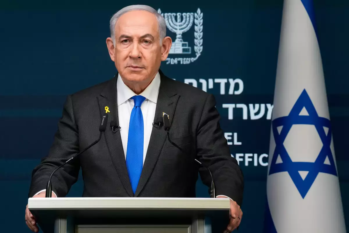 Benjamin Netanyahu, un hombre blanco de 74 años. Cabello blanco canoso, viste un traje negro, una camisa blanca y corbata azul vibrante. Porta una expresión seria. Detrás de él, una bandera de Israel.