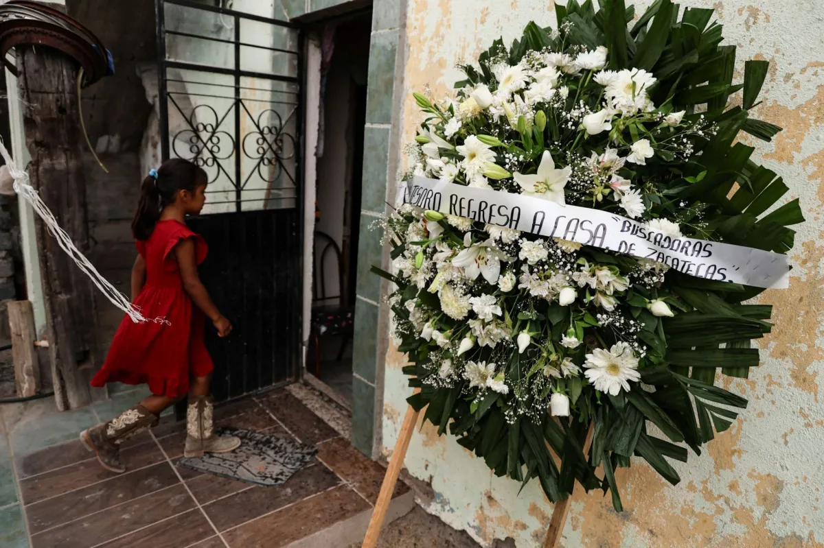 Niña con vestido rojo y botas camina frente a una casa junto a una corona funeraria con un mensaje de familiares de personas desaparecidas.
