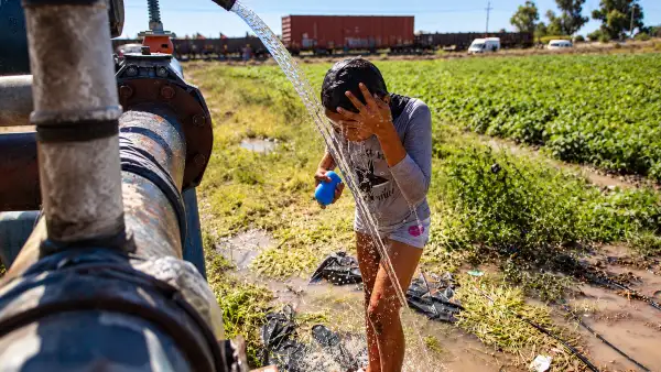 Niña se moja con agua que sale de una tubería en un campo agrícola, mientras un tren de carga circula al fondo.