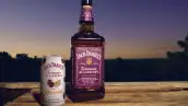 Botella de Jack Daniel’s Tennessee Blackberry y lata de Jack Daniel’s Blackberry Lemonade sobre una superficie de madera, con un paisaje arbolado y el cielo al atardecer como fondo.
