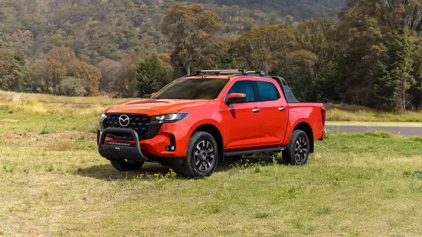 Imagen de la nueva pickup Mazda Bt-50 color rojo, estacionada en un paraje con pasto verde con un bosque y montañas de fondo.