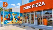 Fachada de un restaurante Dodo Pizza con letrero naranja, ventanales de cristal y un mural colorido en el exterior. Frente al establecimiento hay espacios de estacionamiento y un anuncio vertical con el logotipo de la cadena.