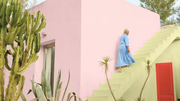 Una mujer asciende por unas escaleras en un edificio pintado con color plasma, un tono rosado. Al frente se observan unas plantas.