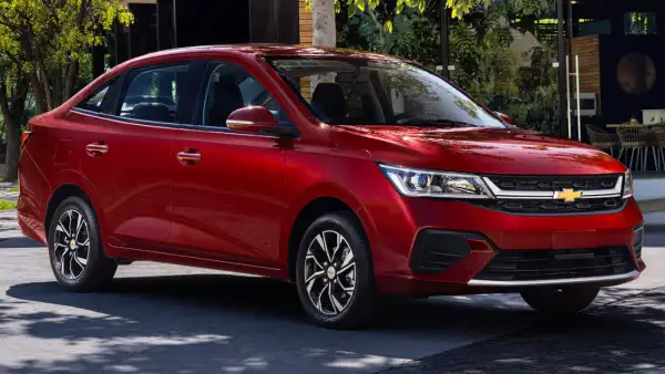 Chevrolet Aveo Sedán 2026 color rojo, visto desde un ángulo frontal de tres cuartos, con parrilla negra, faros modernos y rines de aleación, estacionado en una calle arbolada.