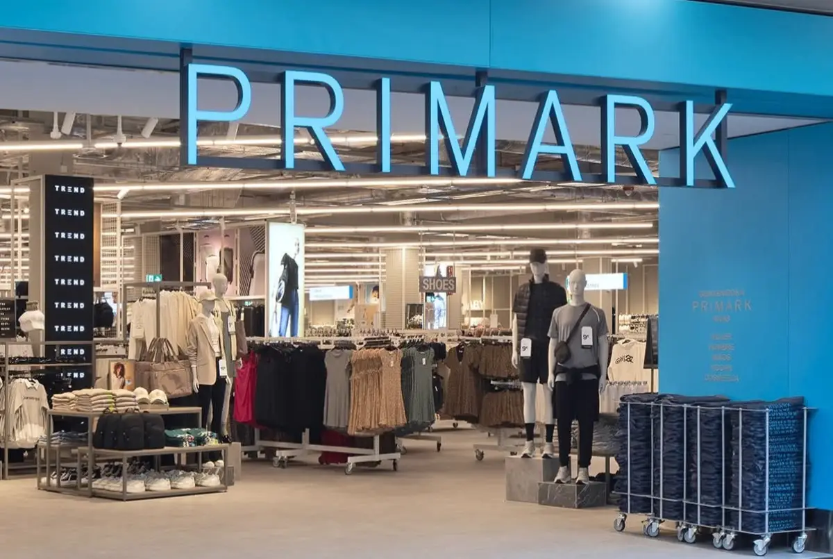 Entrada de una tienda Primark, en la que se muestran aparadores con ropa y zapatos de colores neutros como gris y nude. La fachada es color azul, con el nombre de la cadena en el mismo color en la parte de arriba de la entrada.

