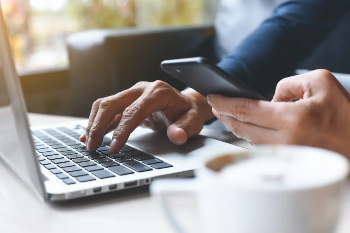 Cierre de un hombre casual o trabajador independiente trabajando en una laptop, sosteniendo un teléfono móvil inteligente con café en una cafetería 