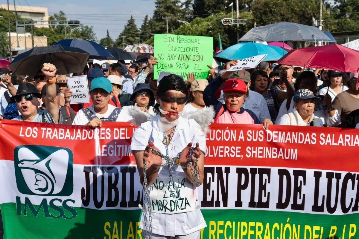 Trabajadores del IMSS protestando en una calle, varios sostienen una manta larga en la que exigen solución a sus demandas salariales; enfrente una persona con el uniforme blanco, las manos amarradas y la boca y los ojos tapados. 