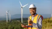 Engineer India man working with tablet at windmill farm Generating electricity clean energy. Wind turbine farm generator by alternative green energy. Asian engineer checking control electric power