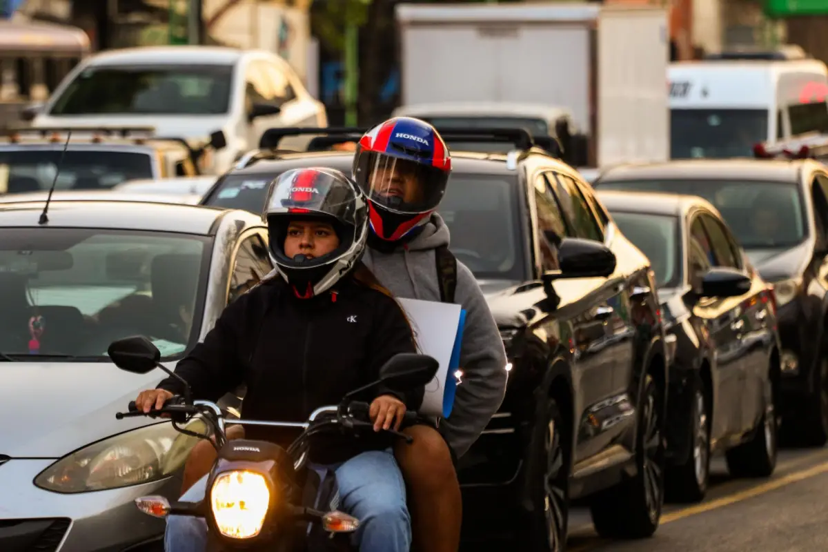 Dos personas viajan en una moto, con otros vehículos de fondo.