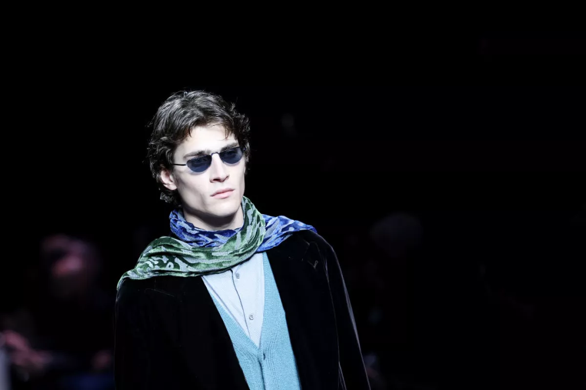 Foto de un modelo en la pasarela de Armani en Milan otoño/invierno 2024-2025 con saco negro de terciopelo, lentes oscuros y una mascada de seda en tonos verdes y azules.