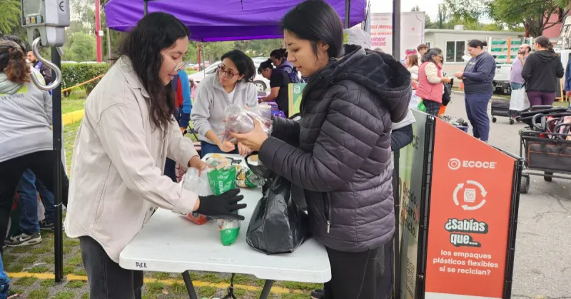 Dos mujeres frente a frente en una carpa al aire libre, una revisa con guantes un recipiente. Otra con chamarra negra lo entrega.jpg