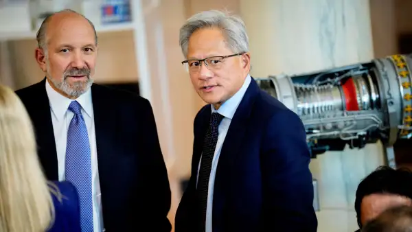 Jensen Huang aparece vestido con un traje azul marino y corbata con puntos durante una reunión. Detrás de él, un aparato tecnológico.