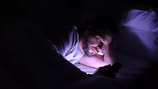 Foto de stock de un hombre acostado en su cama. en un cuarto completamente oscuro y únicamente ilumina su cara el celular