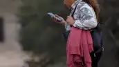 Una mujer en la calle caminando mientras mira su celular con la mano.