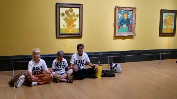 Foto de tres activistas de Just Stop Oil sentados en el suelo y tomados de la mano dentro de un museo en Filadelfia frente al cuadro de Vincent Van Gogh de los 'Girasoles'.