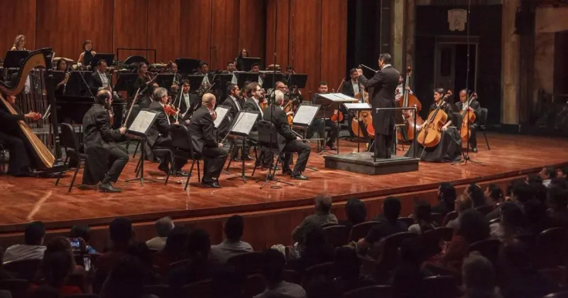 Músicos con violines y partituras con un director de orquesta en un teatro de color café frente a personas sentadasjpg