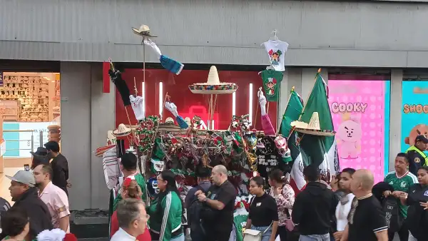 En la imagen se ve un carrito de venta de banderas, mientras personas caminan frente a este. 