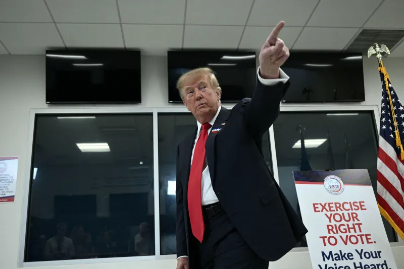 Donald Trump, con traje oscuro y corbata roja, levanta el brazo y señala hacia arriba dentro de un centro de votación en Florida. A su lado hay un cartel que invita a ejercer el derecho al voto y una bandera de Estados Unidos.