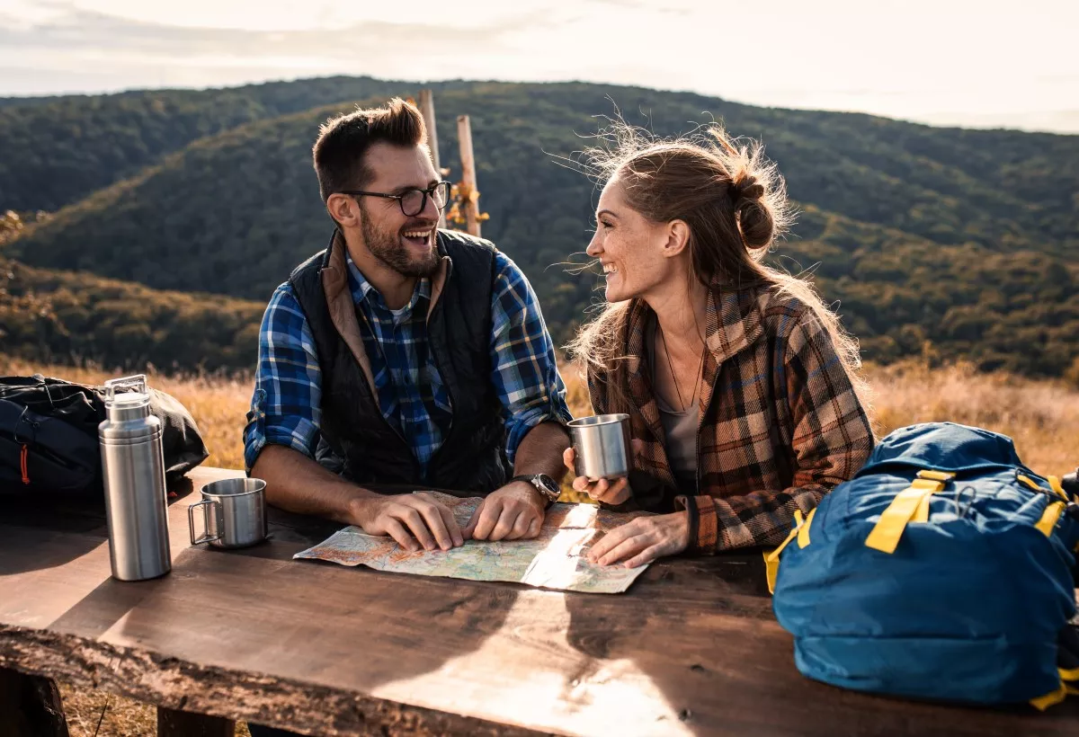 Una pareja está en un bosque disfrutando de la naturaleza, tomando café, con su mochila de campamento y un mapa.