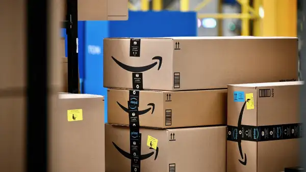 Paquetes de cartón de Amazon, algunos con cinta de embalaje de Prime y etiquetas de envío, apilados sobre una banda transportadora dentro de un centro logístico, con estructuras y maquinaria industrial al fondo.