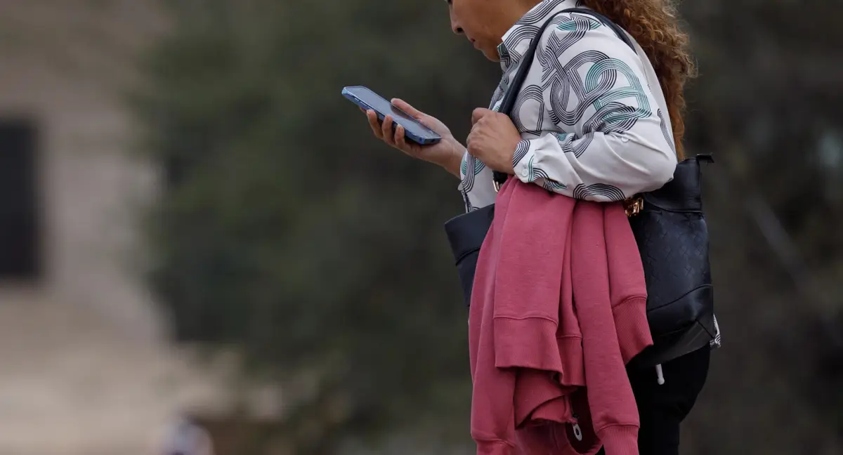 Una mujer en la calle caminando mientras mira su celular con la mano.